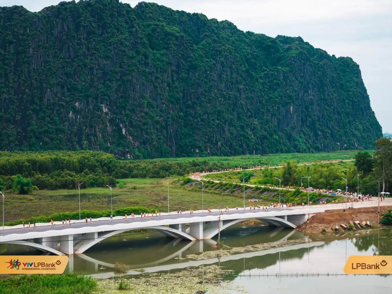 A long line of runners across a bridge and dyke road at the foot of the mountains