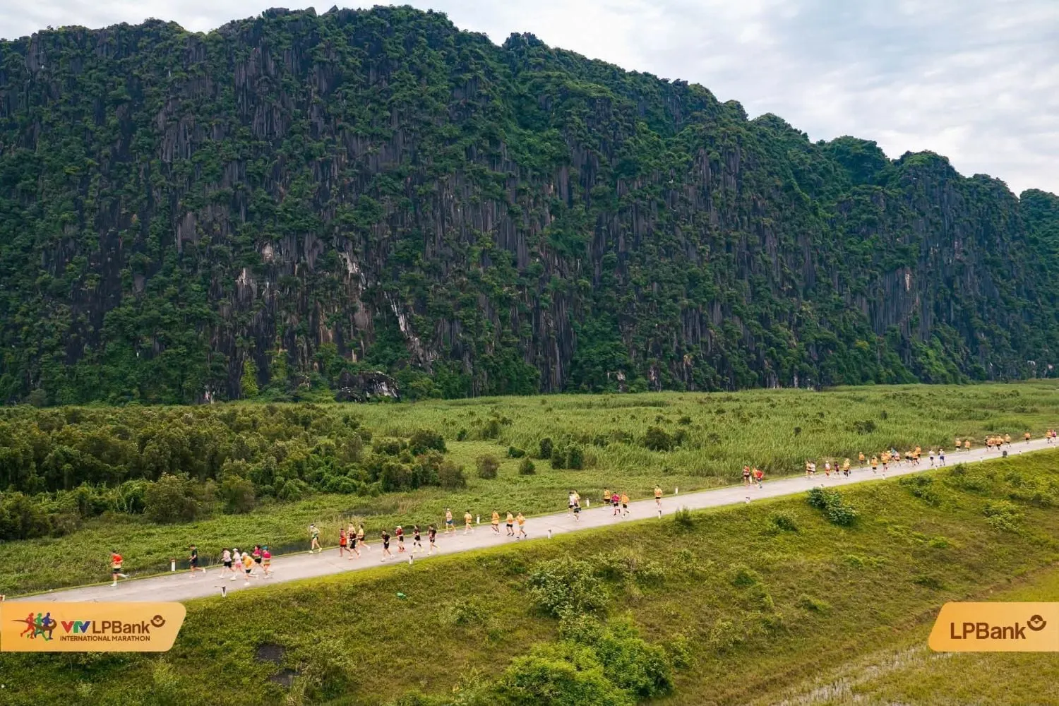 Runners on a dyke road with forested limestone cliffs behind, aerial view